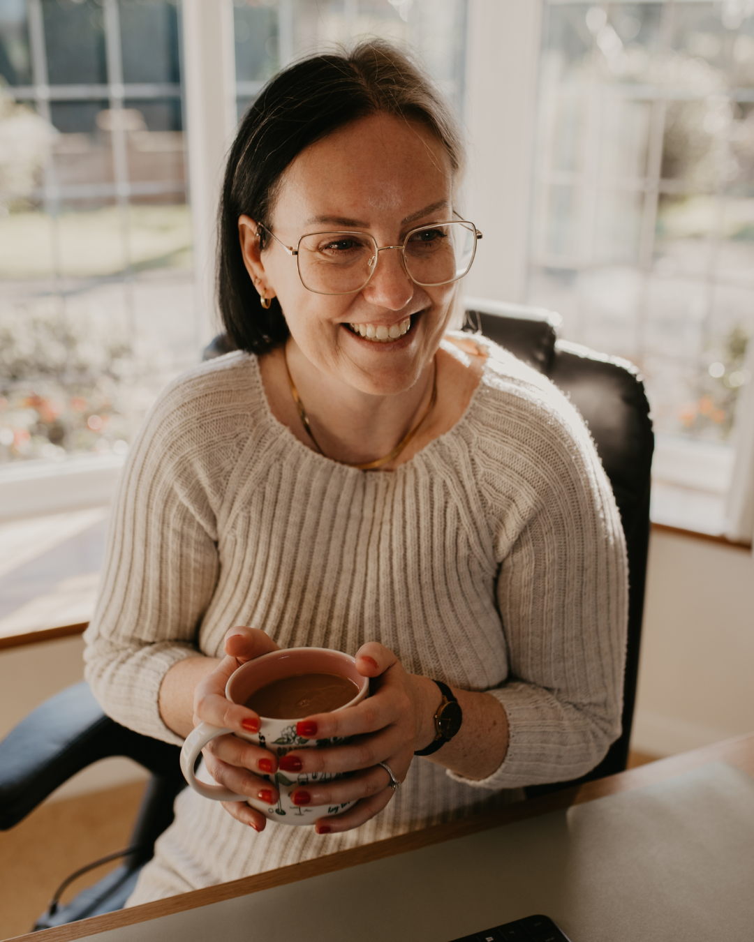 Lady smiling holding a cup of tea. Cream jumper, dark shoulder length hair and wearing glasses