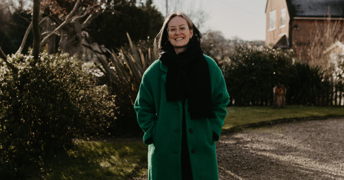 Woman outside with trees, green long coat, black scarf, smiling